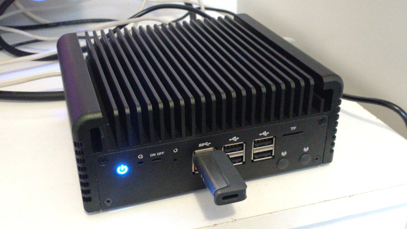 The CWWK fanless mini PC: a solid looking black case with fins along the top to form a heat sink. On the front of the case are six USB-A ports, one of which has a thumb-drive plugged in to it. On the left the power button glows with a blue power logo.