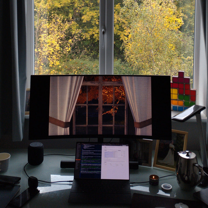 My desk with a laptop and screen; on the screen is a scene from Draugen showing a view out of the window beyond which a silver birch with orange leaves; behind my monitor the view out of my window showing trees with autumnal leaves as well (more yellow than orange).