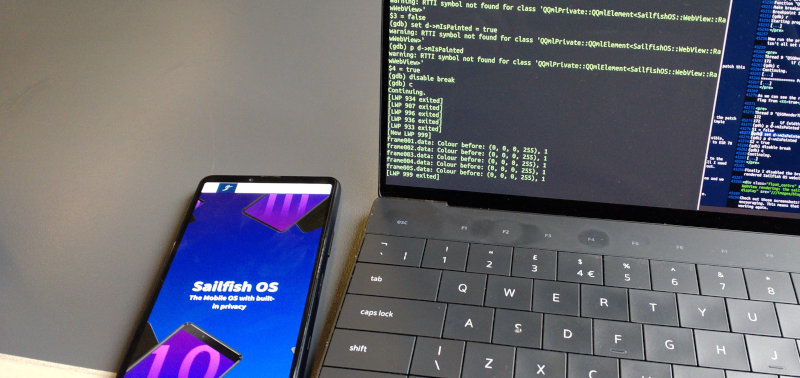 A phone showing the sailfishos.org website next to a laptop on a table with the debugger running in a console window.
