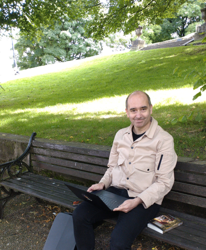 Me, sitting on a park bench in The Slopes, Buxton, with a laptop, a phone and a book. The sun is shining and in the background a grassy hill stretches above me.